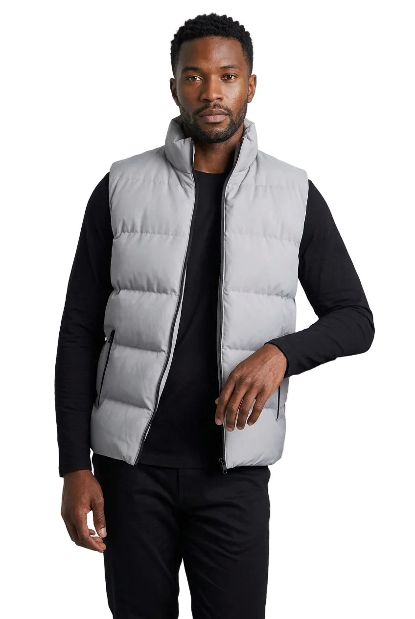 A man wearing a light gray zip-up puffer vest over a black long-sleeve shirt and black pants, standing in a studio setting next to a gray block.