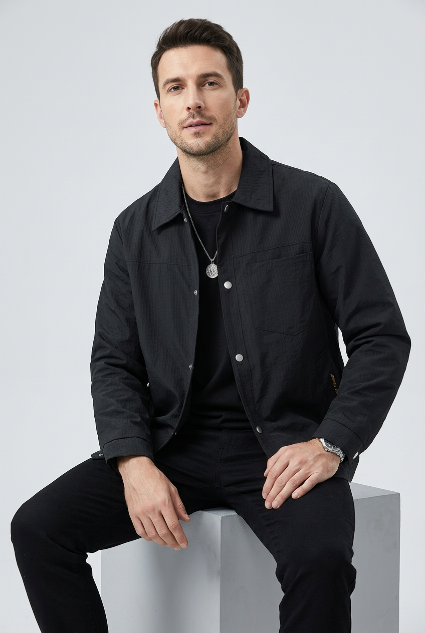 Slim male model wearing REEVEAR black snap-down jacket, seated pose